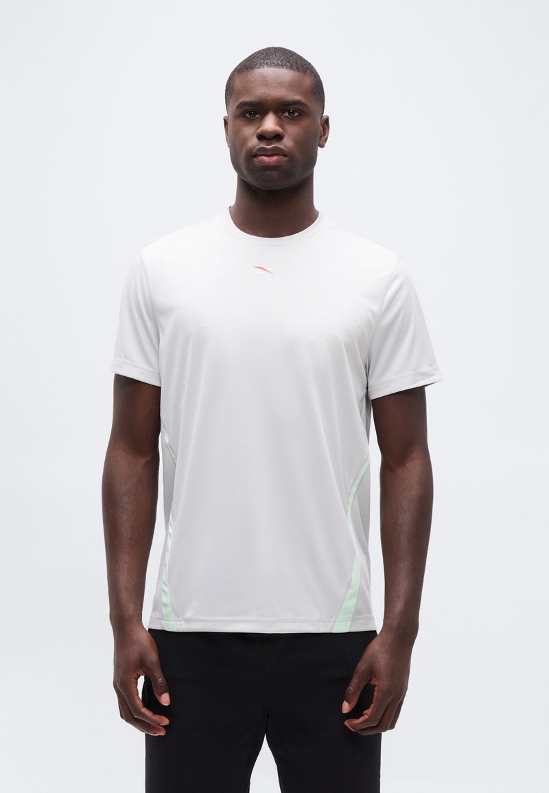 Άνδρας που στέκεται εξερχόμενος προς τα εμπρός φορώντας λευκό αθλητικό μπλουζάκι με κοντά μανίκια, ανοιχτοπράσινες πλαϊνές λωρίδες και μαύρο σορτς, μπροστά σε λευκό φόντο.