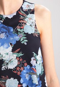 Top senza maniche con fantasia floreale su sfondo nero; presenta grandi fiori blu e bianchi, foglie verdi e piccoli accenti rossi.