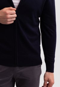 Námořnický svetr na zip vyrobený z jemného pleteného materiálu. Má dlouhé rukávy a žebrovaný lem. Ruka drží zip blízko límce.