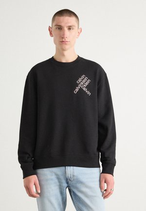 Black cotton sweatshirt with a crew neck, featuring diagonal white text "calvin klein" on the left chest. Casual fit and long sleeves.