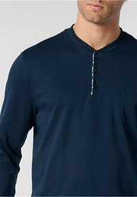 Maglietta a maniche lunghe blu navy con scollatura rotonda, patta con tre bottoni e taschino sul lato sinistro del petto. Tessuto liscio con una leggera trama.