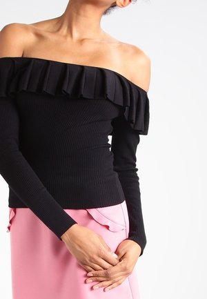 Femme portant un top noir côtelé à épaules dénudées avec un col à volants et des manches longues, associé à une jupe rose avec un détail volanté.