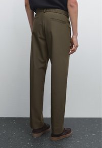 Pantalons décontractés de couleur vert olive, avec une taille élastique, des jambes fuselées, une poche arrière unique, portés avec des chaussures marron foncé.