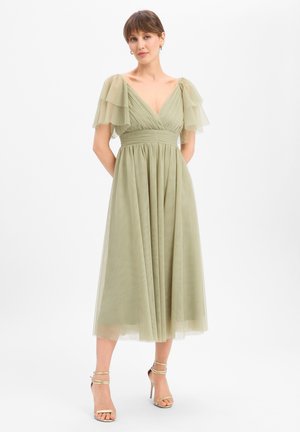 Femme portant une robe midi vert clair avec des manches bouffantes superposées, un col en V, une taille cintrée et des sandales à talons hauts à brides sur un fond uni.