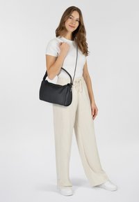 Bolso de mano de cuero negro con una textura suave, forma curva y correa ajustable. El modelo lo lleva con pantalones ligeros y holgados y zapatillas.