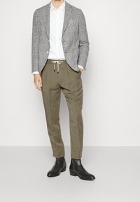 Men's outfit featuring a gray checked blazer, white shirt, olive green pleated pants, and black ankle boots. Contrast detailing on pants.