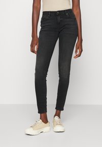 Jeans negros y ajustados con cintura de altura media, que presentan un diseño estándar de cinco bolsillos y un leve desvanecimiento. Combinados con zapatillas beige.