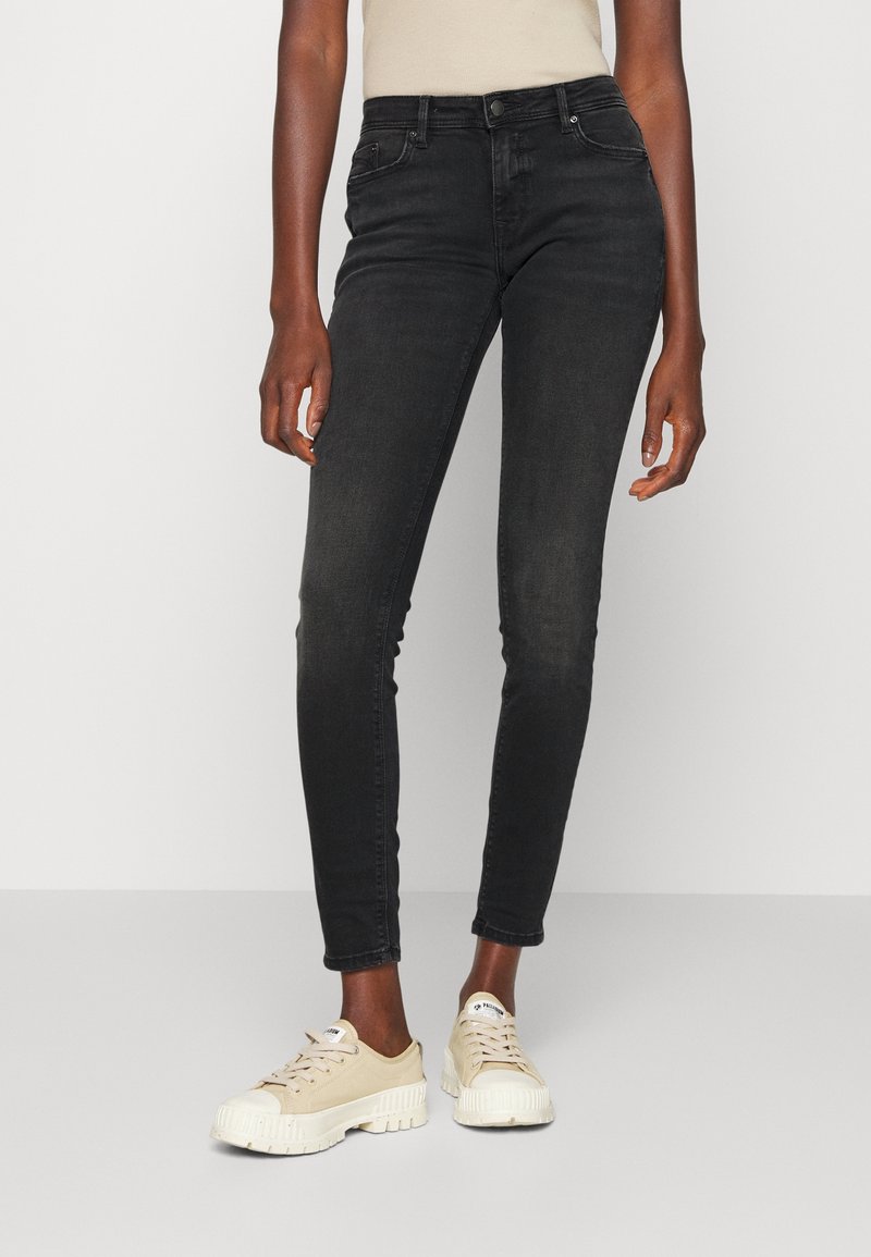 Jeans negros y ajustados con cintura de altura media, que presentan un diseño estándar de cinco bolsillos y un leve desvanecimiento. Combinados con zapatillas beige.