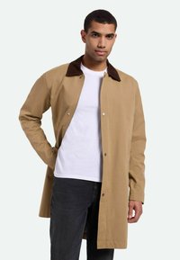 Beige Mantel mit einem braunen Cordkragen, ausgestattet mit einem Frontreißverschluss, Druckknöpfen und langen Ärmeln, getragen über einem weißen T-Shirt und schwarzen Jeans.