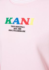 Camiseta rosa con el texto multicolor "KANI" en verde, azul, rojo y amarillo, con texto negro debajo que dice "THE ORIGINAL EST. 1989 REG. TRADEMARK."