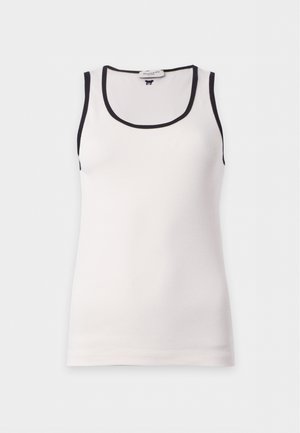 Frau steht, trägt ein weißes Tanktop mit schwarzem Besatz, khakifarbene Shorts und schwarze Knöchelstiefel, mit einer Hand in der Tasche.