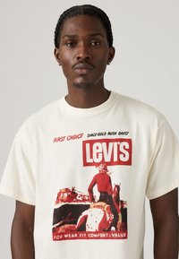 Witte katoenen t-shirt met een rode grafische print van een cowboy en paard, met het "Levi's" logo en tekst in zwart, wat het vintage ontwerp benadrukt.