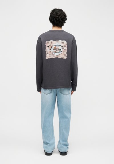 Άνδρας με σγουρά μαλλιά φοράει σκούρο μακρυμάνικο πουκάμισο με ανάμεικτη γραφική στάμπα στην πλάτη, συνδυασμένο με ανοιχτό μπλε τζιν και μαύρα παπούτσια, γυρισμένος από πίσω.