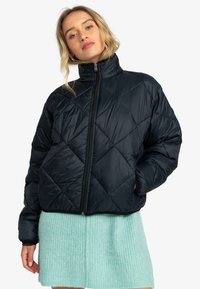 Navy blue quilted puffer jacket with a high collar, zip closure, and a shiny texture, paired with a mint green textured skirt.