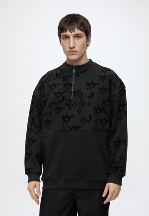 Junger Mann trägt einen schwarz gemusterten Half-Zip-Pullover mit langen Ärmeln und dunkler Hose, steht vor weißem Hintergrund.