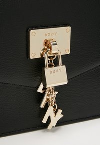 Borsa a tracolla in pelle nera con una targhetta dorata DKNY, lucchetto e charm decorativi su una catena. Texture liscia con un design strutturato.