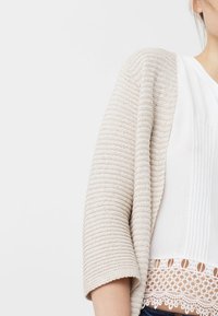 Beige pletený cardigan s texturovaným vzorem a širokými rukávy, přes bílé tričko s sklady a krajkovým lemem.