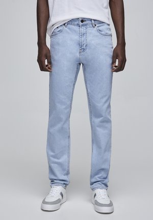 Man wearing light blue jeans, white sneakers with gray details, and a white t-shirt standing against a plain background.