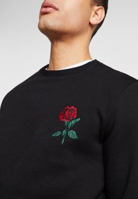 Felpa nera in cotone, con un design ricamato di una rosa rossa e foglie verdi nella parte sinistra del petto. Collo rotondo.