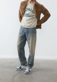 Hombre con chaqueta de pana marrón, sudadera blanca con el texto "Dinamarca Copenhague", vaqueros desgastados de color azul y zapatillas azul y blanca sobre un suelo gris.