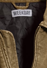 Gros plan d'une veste marron ouverte avec une étiquette blanche visible à l'intérieur portant la mention "WEEKDAY" et une fermeture éclair partiellement ouverte.