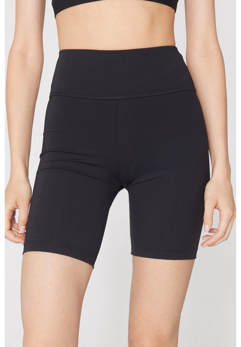 Frau trägt schwarze, hoch taillierte, enganliegende Shorts und ein schwarzes Crop-Top, steht mit entspannten Händen an den Seiten.