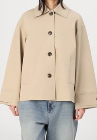 Beige, oversized jakke lavet af bomuldsstof. Har en klassisk krave, knaplukning og to sidelommer. Blå denimbukser under.