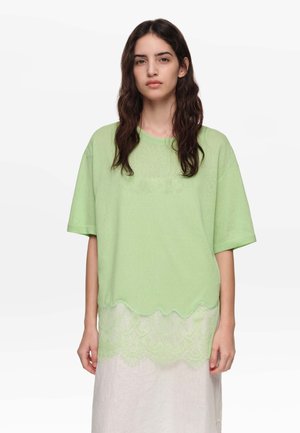 Vrouw met lang donker haar, gekleed in een lichtgroen oversized T-shirt met kanten detail aan de zoom en een witte rok tegen een effen achtergrond.