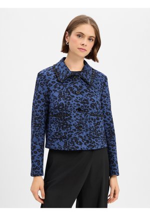 Femme portant une veste boutonnée à imprimé léopard bleu et noir avec col et un pantalon noir, debout devant un fond blanc uni.