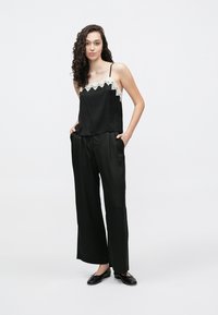 Black satin camisole with lace trim at the neckline, paired with loose black pants. Model stands on a plain background. Balanced silhouette.