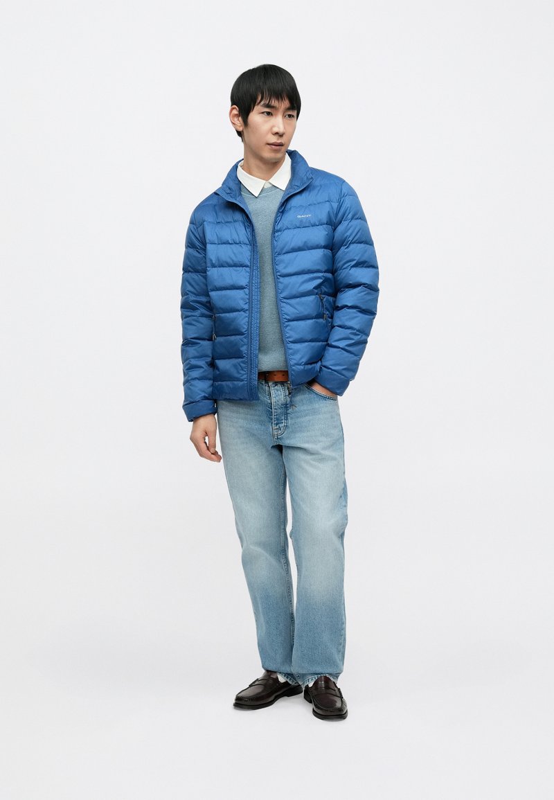 Blue puffer jacket with a zip front, horizontal quilted design, and high collar. Worn over a light blue sweater and paired with light jeans.