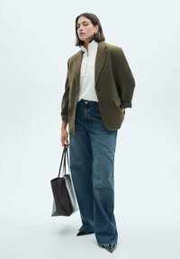 Olivovo zelený oversize blazer, biely zip-up top, modré široké nohavice, čierne lodičky na špicatú špičku a čierna kabelka. Jednoduchý, štruktúrovaný dizajn.