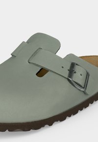 Κοντινό πλάνο ενός γκρι σανδαλιού slip-on με ασημένια πόρπη και καφέ υφή σόλας σε λευκό φόντο.