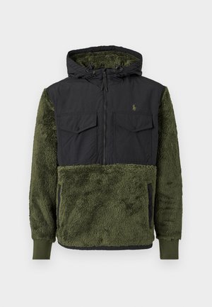 Mēteļa jaka ar kapuci, kurai augšējā daļa ir melna, bet apakšējā daļa ir zaļa, teksturēta vilna. Iekļauj priekšējās kabatas un rāvējslēdzēju.