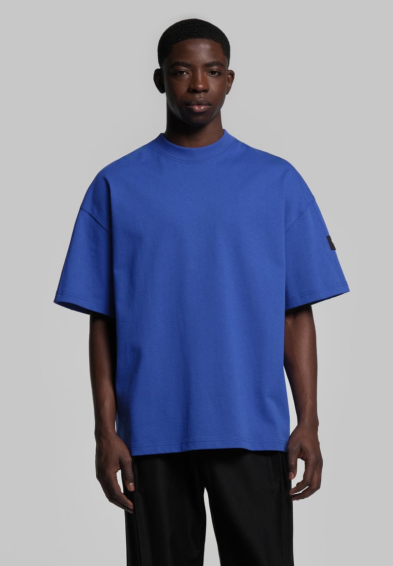 T-shirt bleu surdimensionné à manches courtes, fabriqué en coton doux, avec un col rond côtelé et une petite étiquette noire sur la manche gauche.