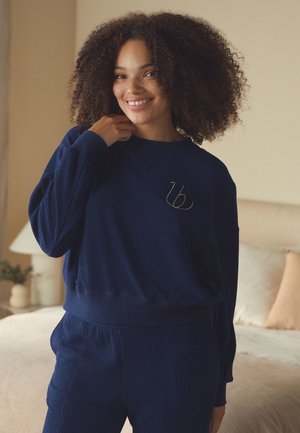 Usmievajúca sa žena s kučeravými vlasmi v tmavomodrej textúrovanej mikine a zodpovedajúcich nohaviciach stojaca v jemne osvetlenej spálni.