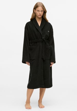 Frau steht barfuß in einem langen schwarzen Bademantel aus Frottee mit Taschen und einem gebundenen Gürtel vor einem schlichten weißen Hintergrund.
