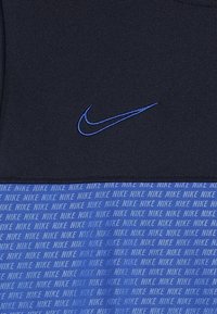 Nike sportkleding met een donkerblauwe bovenkant met een blauwe geborduurde logo, en een ondergedeelte in blauw met een herhaald "NIKE" tekstpatroon.