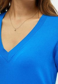 Pull bleu à col en V en tissu tricoté, avec des bords côtelés. Une délicate chaîne en or avec un pendentif en forme de cœur est également visible.