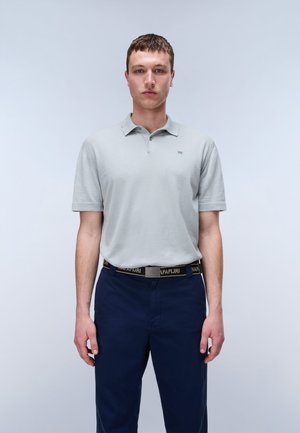 Junger Mann steht mit Blick nach vorne, trägt ein hellgraues kurzärmeliges Poloshirt und marineblaue Hose mit schwarzem Marken-Gürtel.