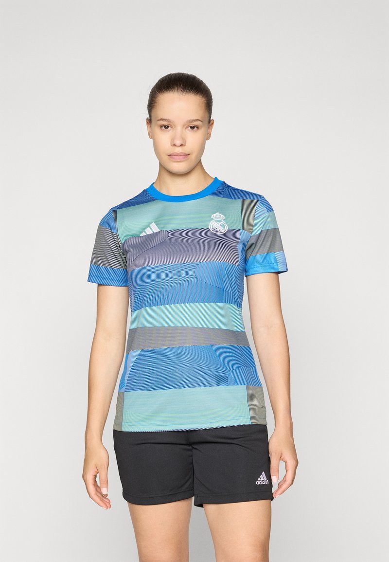 Kortærmet blå sportsbluse med bølgende mønstre, prydet med Real Madrid-logoet og Adidas-striber, som er sat sammen med sorte shorts.