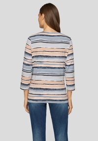 Eine Frau trägt ein gestreiftes Langarmshirt in Blau-, Beige- und Schwarztönen, kombiniert mit blauen Jeans, von hinten vor einem grauen Hintergrund gezeigt.