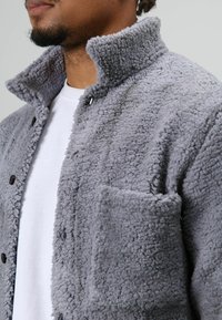 Grijze fleecejas met een hoge kraag, knoopsluiting en een borstzak. Zachte textuur met een weelderige, gezellige uitstraling.