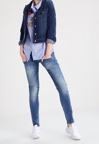 Jeansowa kurtka z podwiniętymi rękawami, nałożona na jasnoniebieską koszulę w paski. Dopasowane niebieskie dżinsy i białe trampki dopełniają wygląd.