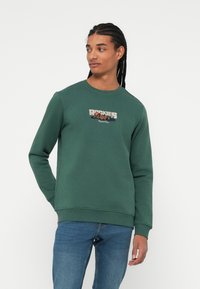 Groene sweatshirt met lange mouwen, ronde halslijn en ribgebreide manchetten. Heeft een "ROCKIES" grafiek in beige en oranje op de borst.