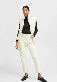Ivoorgekleurde gewatteerde vest met hoge kraag, gedragen over een zwarte lange mouwen top. Gecombineerd met off-white rechte broek en zwarte instappers.