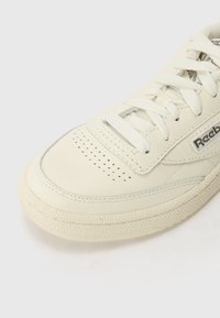 Hvit lær sneakers med perforert tåboks, flate snørebånd og teksturert gummisåle. Har en sølvfarget logo på siden.