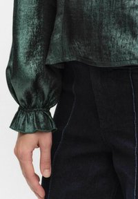 Zielona, półprzezroczysta bluzka o teksturowanej powierzchni, z długimi rękawami i falbaniastymi mankietami. Połączona z ciemnymi dżinsami z niebieskimi szwami.