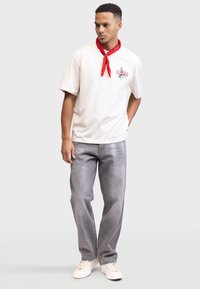 Jasno-beżowy t-shirt z czerwonym graficznym detalem i krótkimi rękawami, zestawiony z szarymi dżinsami i białymi trampkami. Czerwona chustka związana wokół szyi.