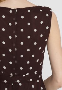 Ruskeasta kankaasta valmistettu mekko, jossa valkoiset pilkut, hihaton malli ja vetoketjukiinnitys takana. Vyötärö on korostettu vyöllä.